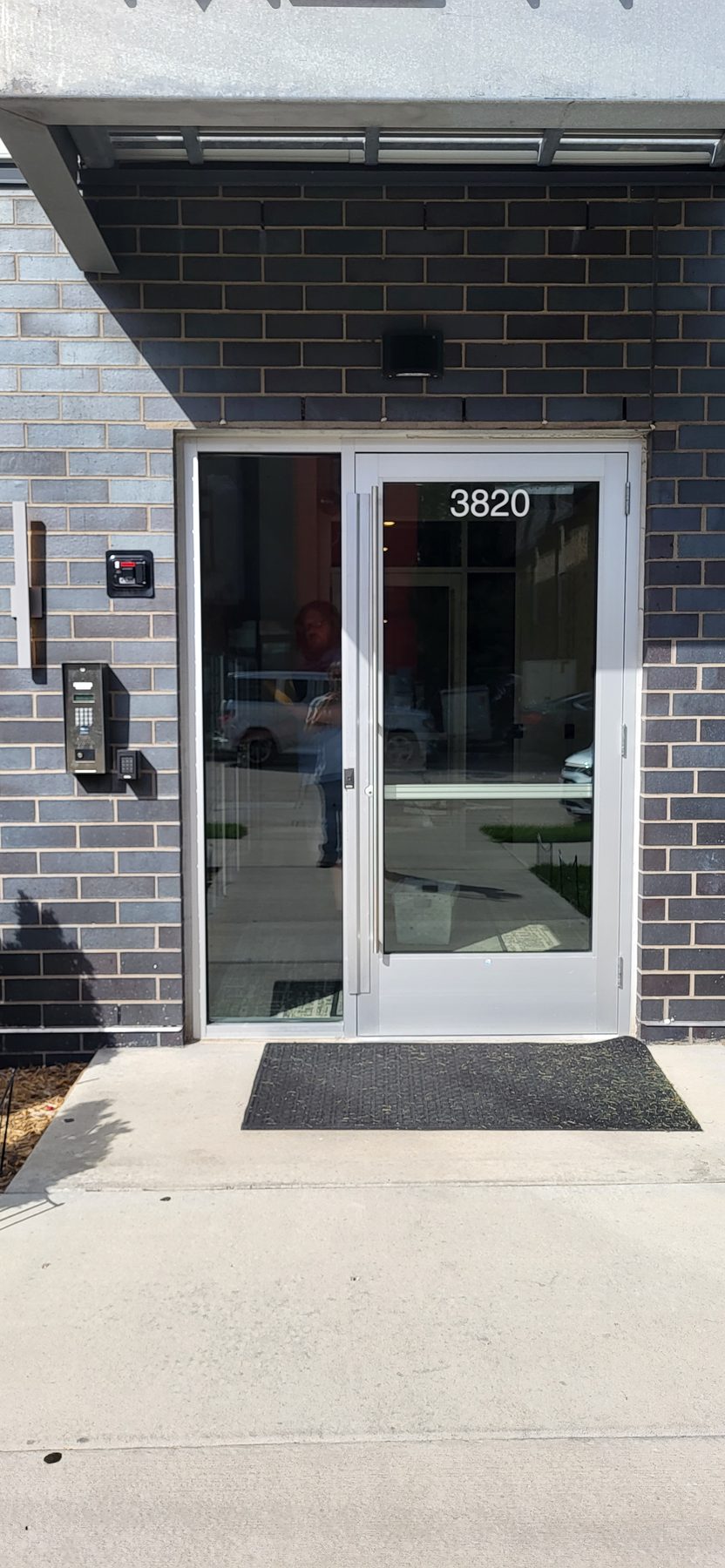 Glass building entrance with an intercom entry panel and card reader mounted on the brick beside the door