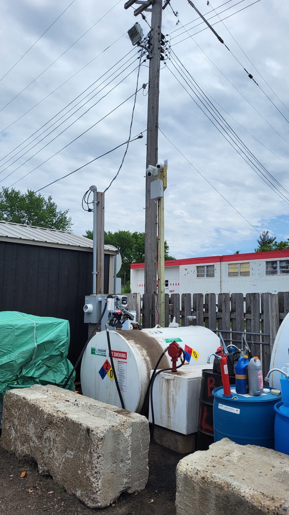Camera and enclosure mounted on a pole beside a fenced fuel storage area in the yard