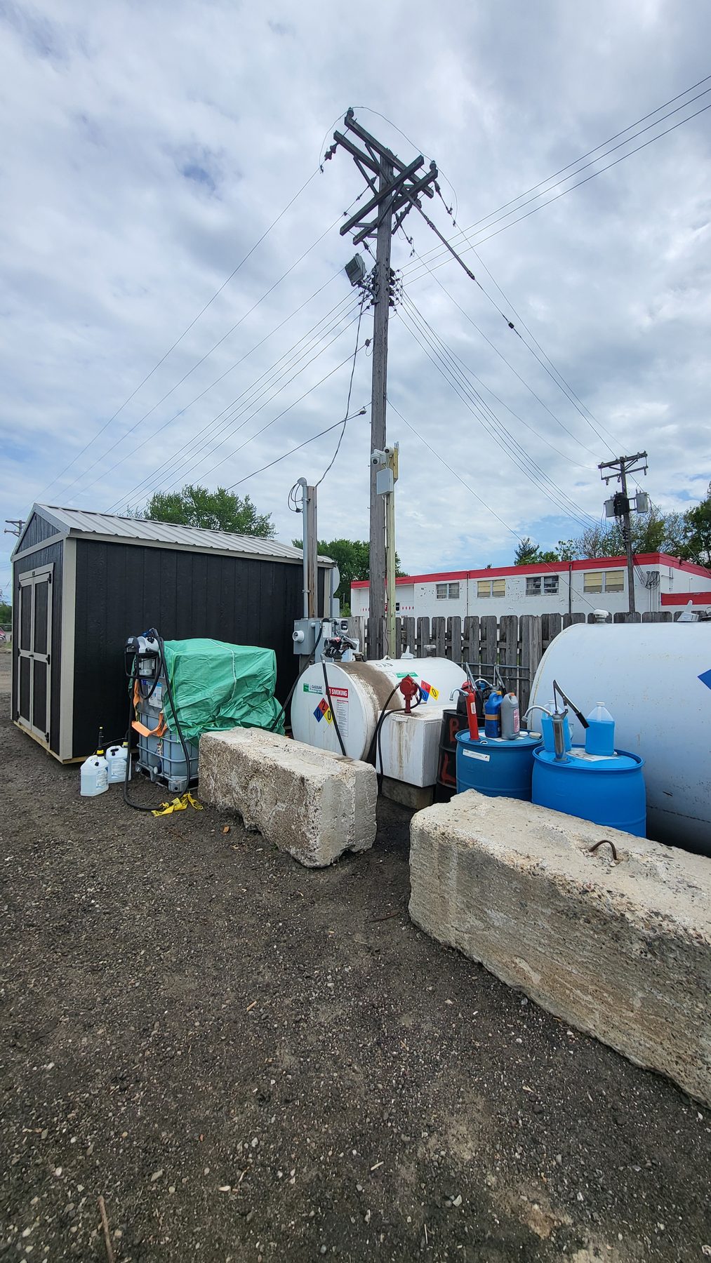 Pole-mounted security camera above an industrial yard with fuel tanks, drums, and a storage shed below